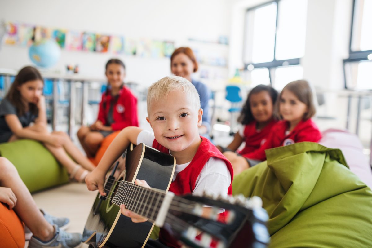 A Down Syndrome Boy With School Kids And Teacher Sitting In Class, Playing Guitar.
