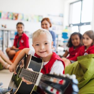 A Down Syndrome Boy With School Kids And Teacher Sitting In Class, Playing Guitar.